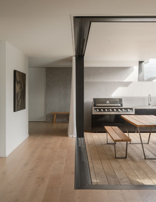 A bright, minimalist open-plan interior featuring a large operable glazed opening with dark metal framing between an indoor hallway/living area and an outdoor entertaining space. The interior has pale timber flooring, white walls, a grey textured feature wall, and a simple timber bench. The adjoining outdoor area has timber decking, a built-in black barbecue kitchen, and a timber dining table with benches. Strong sunlight creates crisp shadows across the pale surfaces.