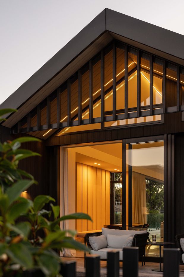A dramatic dusk exterior view of a contemporary dark-clad house with a steep gabled roof, extensive black-framed glazing, warm timber-lined interiors, and integrated linear lighting. The open ground-level glazing reveals a furnished living area and covered outdoor seating, while leafy planting partially frames the foreground.