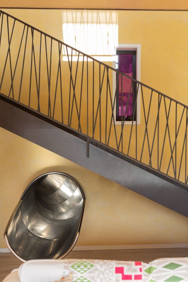 Interior architectural photograph looking upward across a diagonal industrial-style staircase with dark metal balustrades and a broad grey stair stringer. Warm ochre plaster walls, a narrow vertical window with vivid purple glazing, and a polished stainless-steel tube slide are visible, with part of patterned upholstered seating at the bottom edge.