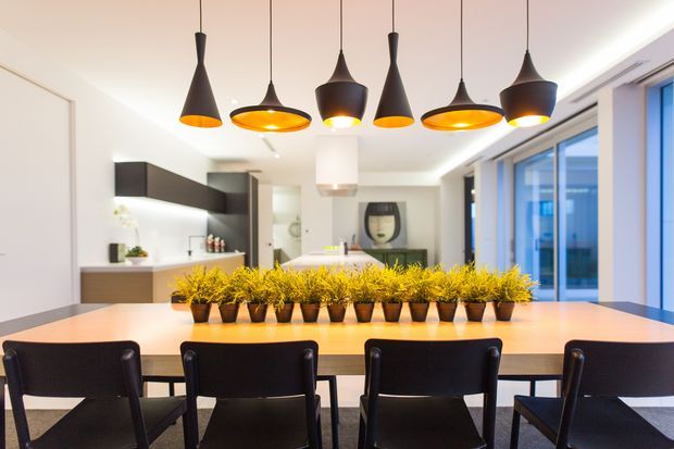 Contemporary open-plan dining area with a long light-wood dining table, black dining chairs, a row of potted plants as a centrepiece, and multiple sculptural pendant lights. A streamlined kitchen with dark cabinetry is visible behind the table, while large glazed doors or windows line the right side.