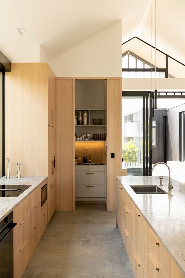 A bright, contemporary kitchen with a central island, pale timber cabinetry, white stone benchtops, integrated appliances, and a compact walk-in pantry or scullery at the rear. The kitchen opens toward a light-filled living area with large black-framed glazing and a high pitched ceiling. A polished concrete floor and linear pendant lights add a refined modern character.