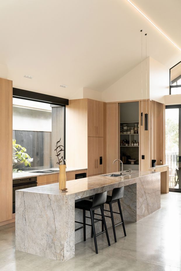 A bright, refined contemporary kitchen with a large full-height stone island and integrated waterfall ends, seating for three on black bar stools, warm timber cabinetry, a concealed pantry, and black-framed glazing. Linear pendant lights and concealed ceiling lighting emphasize the clean open-plan design, while an outdoor courtyard is visible through the windows.
