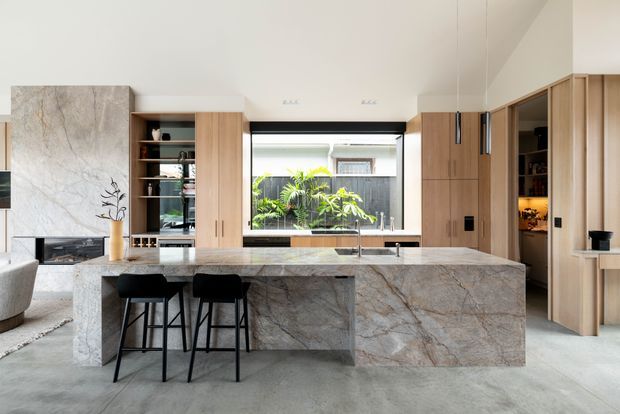 A bright, contemporary open-plan kitchen centered on a large stone island with an integrated waterfall-style end panel and two black bar stools. Full-height light timber cabinetry and open shelving line the walls, with a dramatic veined stone splashback or feature wall behind the island. A large glazed opening provides greenery views, while concealed appliances and warm under-cabinet lighting create a refined, minimalist feel.