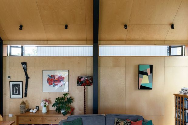 Interior view of a warm, compact living room with extensive light timber wall and ceiling panelling, a dark structural post, high clerestory windows, and a low timber console displaying artwork, ceramics and plants. A grey sofa occupies the foreground, with a bookshelf and personal furnishings at right. The space has a mid-century-influenced, relaxed contemporary character.