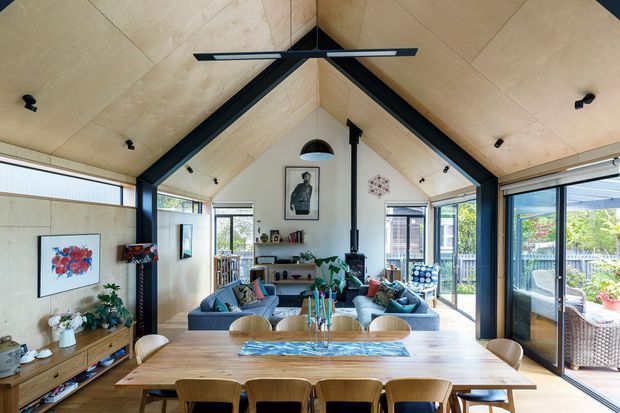 A bright open-plan living and dining room beneath a dramatic pitched, timber-lined ceiling with exposed dark structural beams. The room features a large timber dining table, upholstered dining chairs, a blue sectional seating area, a central fireplace or flue, extensive glazing and sliding doors opening to an outdoor terrace. The interior combines natural timber, black framing, contemporary furnishings and abundant daylight.