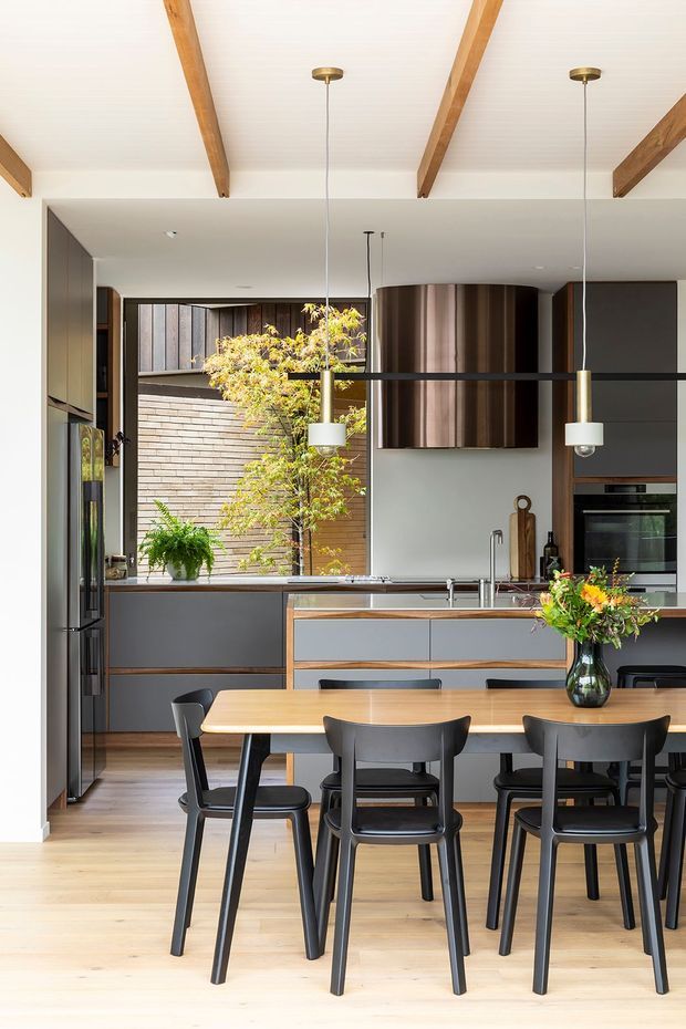 Bright open-plan kitchen and dining area with a timber dining table and black chairs in the foreground. The kitchen features dark grey cabinetry, timber drawer fronts, a large island with integrated sink, stainless appliances, and a curved metallic rangehood. Full-height glazing looks onto a planted courtyard, while exposed timber ceiling beams and pendant lights add warmth.