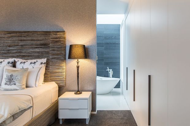 A compact contemporary bedroom with a textured woven headboard, upholstered bed, bedside table and floor lamp. A bright bathroom with a freestanding bathtub is visible through the opening, while full-height white built-in wardrobe cabinetry lines the right side. The room uses a restrained palette of white, grey and natural brown textures.