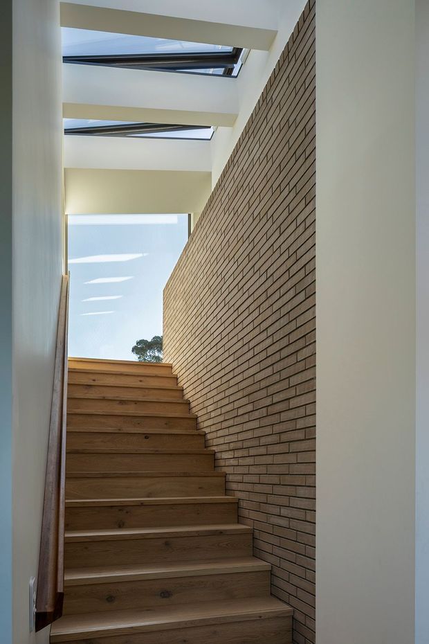 A narrow interior stairway rises beside a full-height exposed brick wall. The stairs have warm timber treads and risers, with white plastered walls and multiple overhead skylights bringing in natural light. An upper landing and a glimpse of trees are visible beyond the stairwell.
