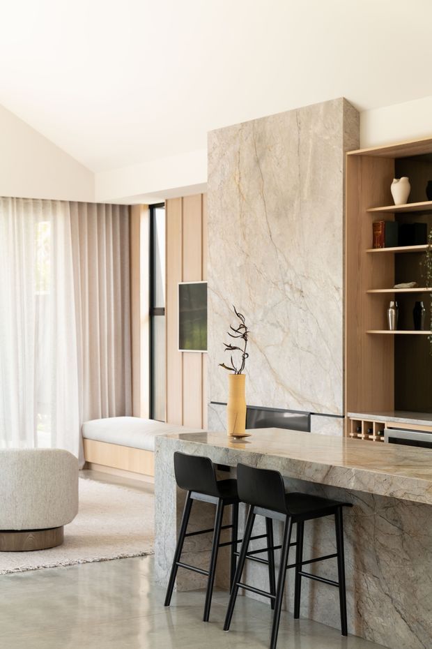 A refined open-plan interior combining a kitchen island and dining counter with a living area. The space features a large pale veined stone feature wall and matching waterfall island, warm timber wall panelling and built-in shelving, black bar stools, a built-in window seat, sheer curtains, and polished concrete flooring. The palette is light, warm, and contemporary with understated natural detailing.