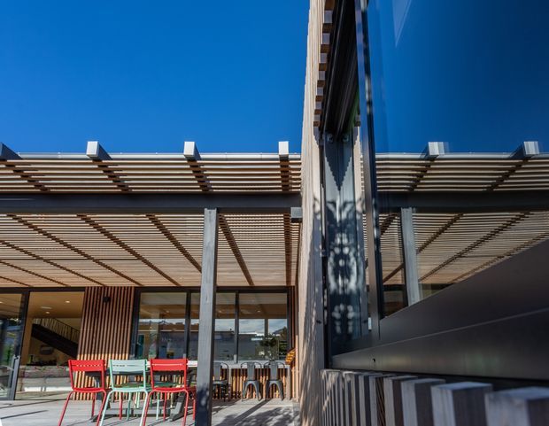 Contemporary commercial or hospitality courtyard viewed from an outdoor dining area, featuring a large glazed facade, reflective vertical glazing, a steel or concrete structural frame, and a slatted timber pergola providing shade. Bright red outdoor chairs and tables occupy the foreground, with a bar or seating area visible behind the glazing.