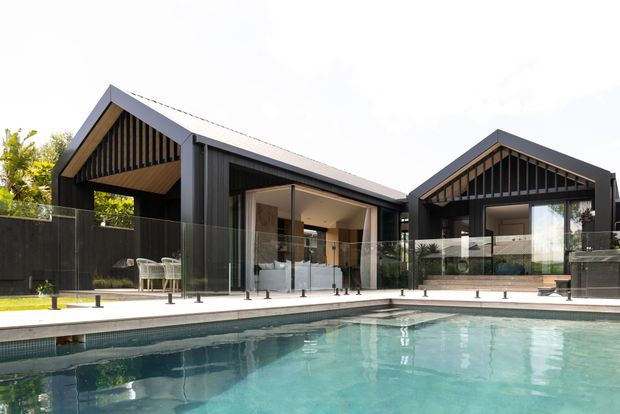 A contemporary dark-clad residential property viewed from a swimming pool. The building comprises two steep gabled pavilion-like forms with dramatic exposed vertical timber detailing beneath the gables, expansive glazing, and a covered outdoor entertaining area opening toward the pool. A glass pool fence and tiled pool surround occupy the foreground.