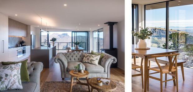 Bright open-plan living and kitchen interior with a tufted grey sofa, timber coffee tables, pale cabinetry, a compact island, and large floor-to-ceiling windows overlooking a mountainous and coastal landscape. A dining area with timber furniture and potted greenery is shown at right. The space has a refined contemporary feel with warm timber flooring and abundant natural light.