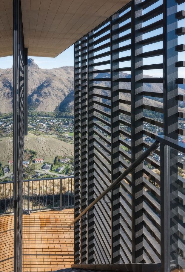 Architectural view from an elevated balcony or terrace, looking across a dramatic mountain valley and hillside settlement. The foreground is defined by a dark vertical louvred screen, black-framed glazing, a timber-toned tiled deck, and a broad projecting soffit. The composition emphasises strong geometric lines, sun-shading elements, transparency, and the rugged alpine landscape beyond.