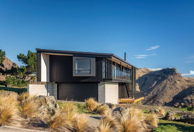 Exterior view of a contemporary elevated mountain house set among rocky terrain and tussock grasses. The building has a dark, rectilinear upper volume with extensive glazing and a balcony, supported by pale masonry or concrete elements, with a low-pitched roof and strong horizontal composition. Dramatic rugged mountains form the background.