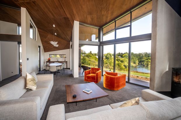 A spacious, contemporary open-plan living room with a double-height timber-lined ceiling, full-height dark-framed glazing, and expansive views toward a leafy waterway. Pale upholstered sofas surround a low dark timber coffee table, while two vivid orange lounge chairs create a strong accent. A dining area, sculptural pendant light, and fireplace are also visible.