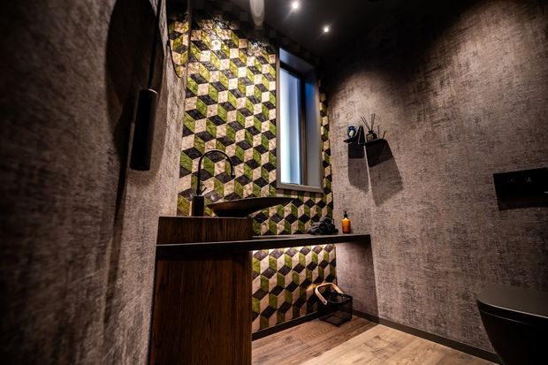 A dramatic, compact contemporary bathroom with dark textured walls, warm timber-look flooring and vanity cabinetry, a black vessel basin, and a freestanding black bath partially visible at right. A bold geometric green, cream and black patterned tile feature wall surrounds the basin and narrow window. Recessed ceiling spotlights, black fixtures, floating shelves and a wall-mounted screen or mirror contribute to the moody, high-contrast design.