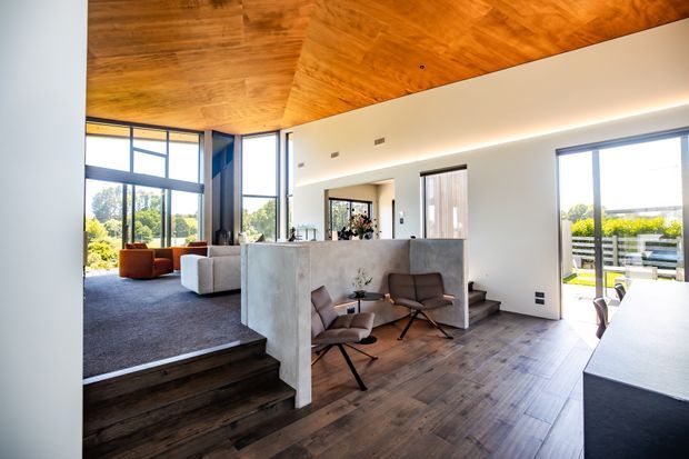 Open-plan contemporary interior with a raised living area, large floor-to-ceiling glazing, a central concrete kitchen island or partition, timber flooring, and a striking warm timber-lined ceiling. The space includes a living room with sofas, a small seating nook, and a dining area visible at right.