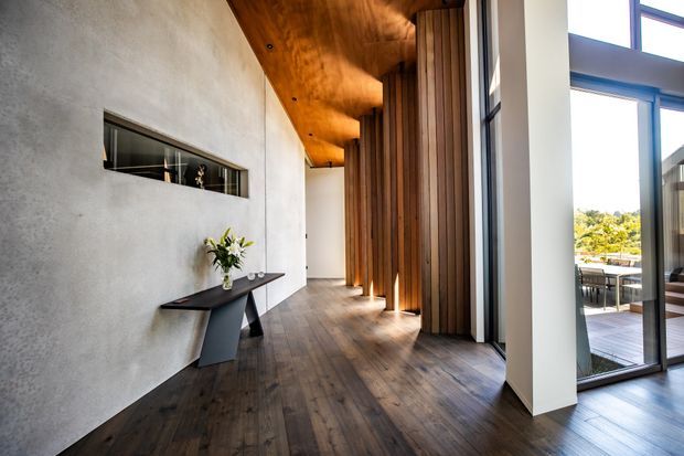 A refined modern interior hallway or entry passage with dark timber flooring, a warm timber-lined ceiling, and a dramatic vertical timber slat feature wall. The left wall has a narrow recessed display niche and a slim sculptural console table, while full-height glazing on the right opens views toward an outdoor deck and landscaped surroundings. The space is long, bright, and carefully detailed with strong natural-material contrast.