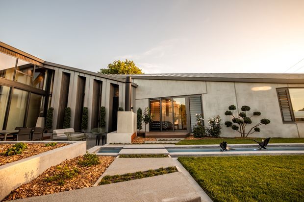 Low, modern residential building viewed from a landscaped courtyard. The composition features a flat-roofed rendered volume beside a darker vertically clad wing with extensive glazing, a paved geometric path, long narrow reflecting pool, manicured lawn, raised garden beds, sculpted shrubs, and outdoor seating. Warm late-day light highlights the contemporary facade and courtyard.