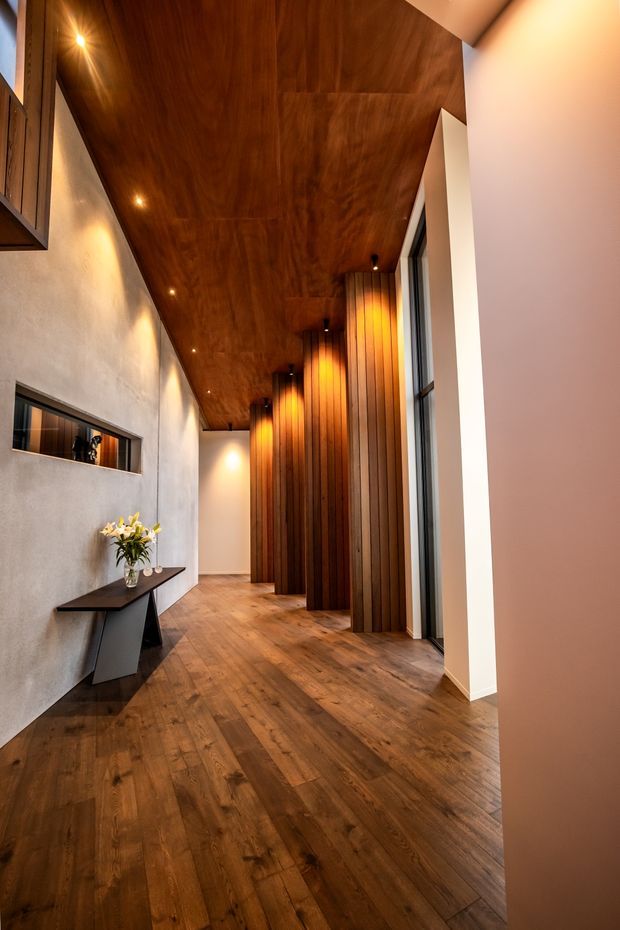 A refined residential hallway or entry passage with a warm timber-lined ceiling, wide timber flooring, and a series of tall vertical timber feature panels concealing or framing doorways along one side. The opposite wall has a textured grey plaster finish, a narrow recessed mirror or opening, and a slim dark console table with flowers. Recessed spotlights and warm wall lighting create an intimate, sophisticated atmosphere, while tall glazing brings in natural light.