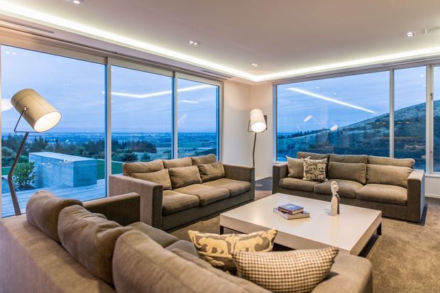 A spacious contemporary living room furnished with multiple taupe sectional sofas, armchairs, a white rectangular coffee table, and floor lamps. Floor-to-ceiling corner glazing provides expansive views over a suburban landscape and hills. Recessed ceiling lighting and a concealed linear cove light create a polished, softly illuminated interior.
