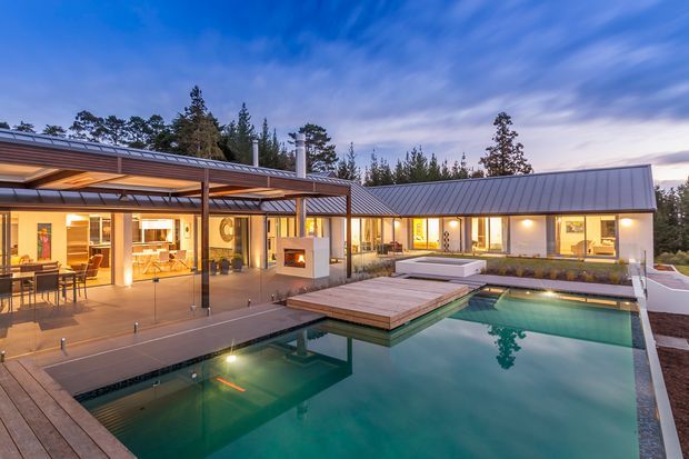 Exterior dusk view of a contemporary single-storey residence arranged around a large swimming pool. The home has a low-pitched metal roof, extensive floor-to-ceiling glazing, a covered outdoor entertaining terrace, timber decking, and illuminated warm interiors visible through the windows. The landscaped setting includes mature trees and a quiet residential outdoor atmosphere.