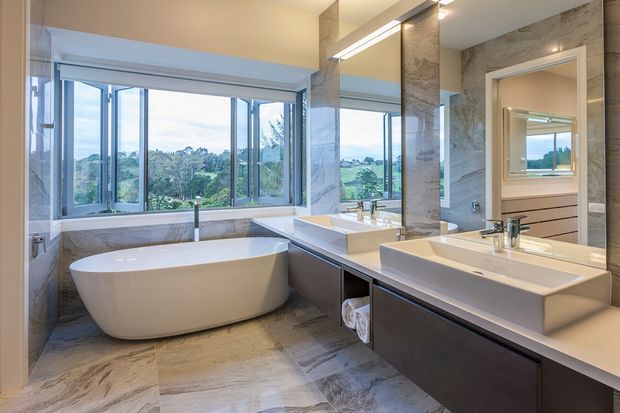 A spacious contemporary bathroom with a freestanding white bathtub positioned beside large open windows overlooking greenery. The room features twin white vessel-style basins on dark built-in vanities, a large wall mirror, warm linear lighting, and grey-beige stone-look wall and floor finishes.