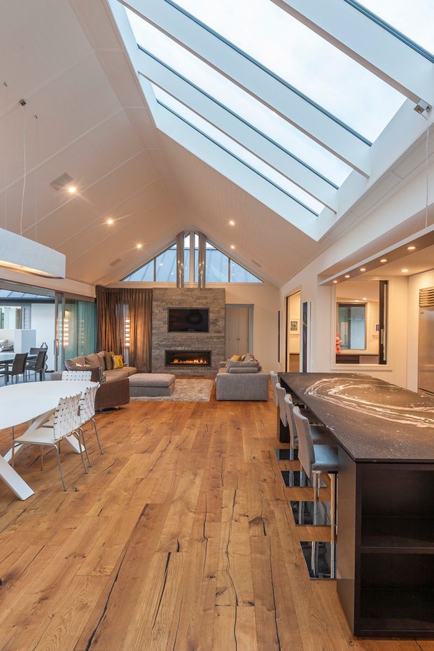 A bright, open-plan contemporary interior combining a living room, dining area and kitchen. The space features a dramatic vaulted ceiling with a large linear skylight, clerestory glazing, exposed warm timber flooring, a stone fireplace wall with built-in television and fireplace, and a large dark kitchen island with seating. Neutral furnishings and extensive glazing create an airy, inviting atmosphere.
