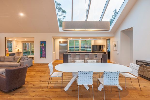 Open-plan contemporary living, dining and kitchen area with warm timber flooring, a large white dining table, modern sling-back chairs, and a charcoal kitchen island. A dramatic linear skylight and suspended pendant provide abundant natural light. The living room is visible to the left, with glazed doors and garden views beyond.