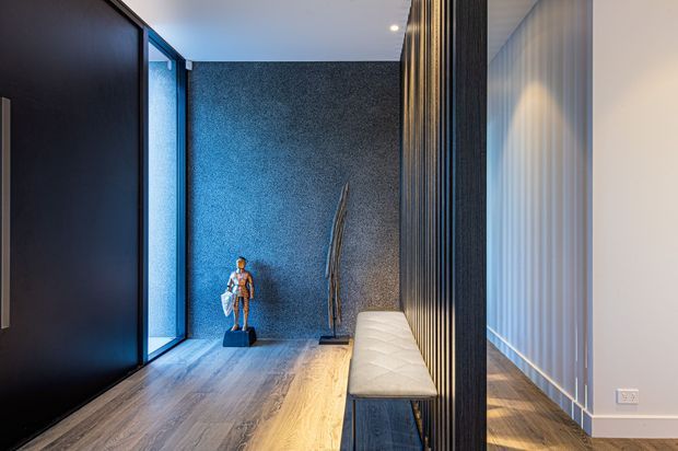 A refined, modern hallway or entry corridor with dark timber or black panelled walls, a textured blue-grey feature wall, floor-to-ceiling glazing, warm timber flooring, and a long upholstered bench beside a vertical slatted screen. Sculptural decor and concealed lighting add a sophisticated, gallery-like atmosphere.