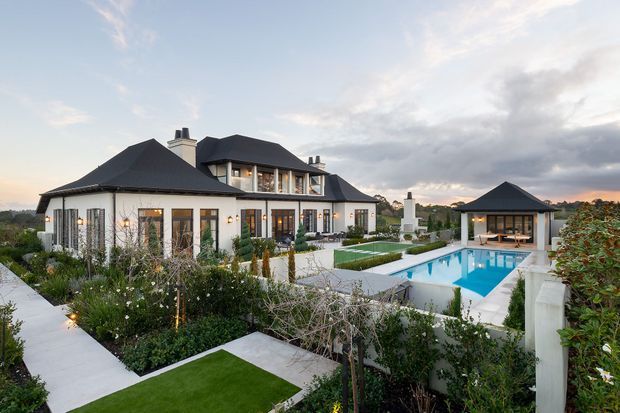A substantial two-storey residence with dark pitched roofs, white rendered walls, extensive glazing, landscaped gardens, a geometric swimming pool, and a separate pool pavilion. The property has formal contemporary-traditional detailing and is photographed at dusk.