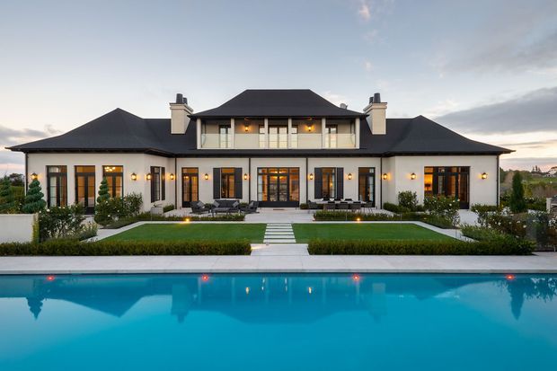 A large symmetrical two-storey residence viewed from across a swimming pool at dusk. The home has white rendered walls, dark hipped roofs, projecting wings, formal window and door arrangements, balconies, chimneys, and extensive glazed openings. A manicured garden with clipped hedges and a central path leads toward the house, while the illuminated pool occupies the foreground.