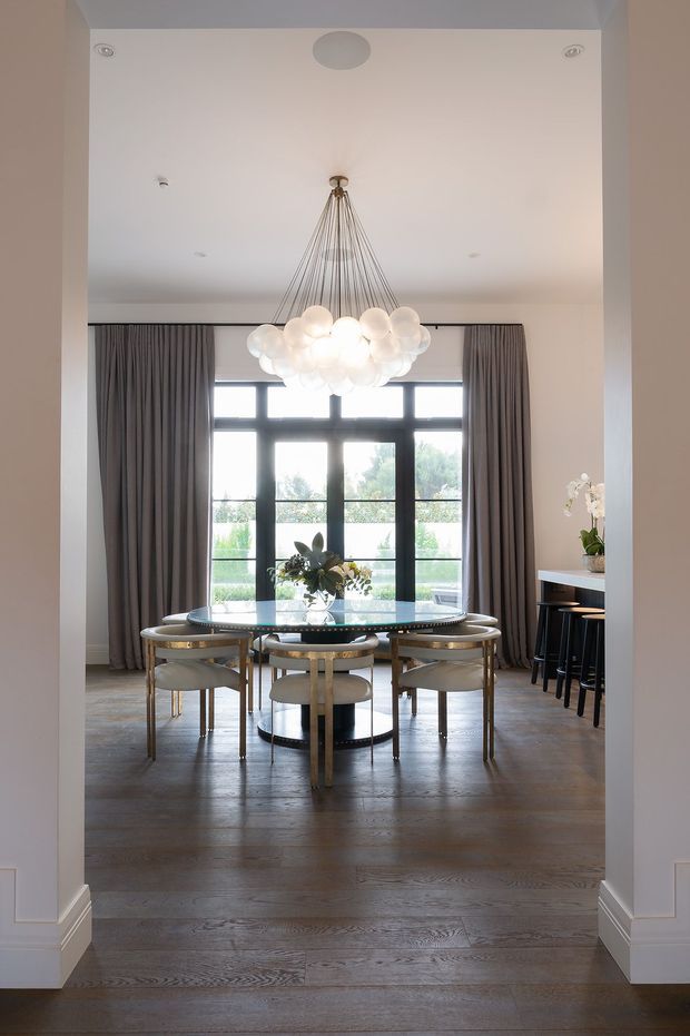 A refined dining room viewed through a wide architectural opening, featuring a round glass dining table with upholstered dining chairs, a sculptural globe chandelier, full-height grey curtains, dark-framed multi-pane windows, and warm timber flooring. A compact bar or serving counter with stools is visible to the right, while greenery is seen outside the windows.