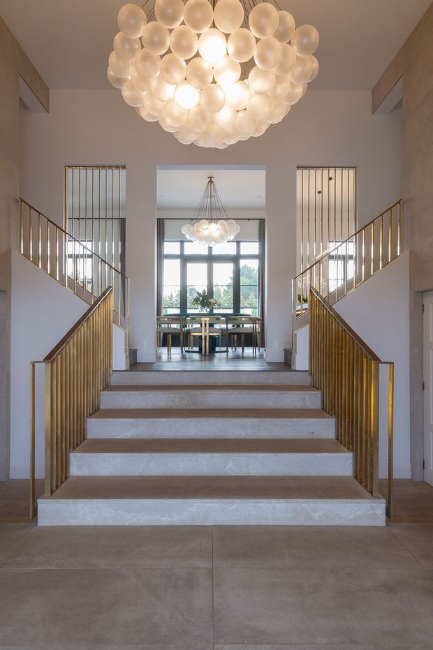 A grand residential stairway rises toward an open dining area, framed by symmetrical white walls and tall vertical windows. The broad stair treads appear to be pale stone or concrete, with warm timber handrails and brass-gold vertical balustrades. A dramatic spherical cluster chandelier dominates the stairwell, while a second matching pendant hangs over the dining table beyond. The interior is bright, refined, and highly symmetrical, with large-format flooring and expansive glazing.