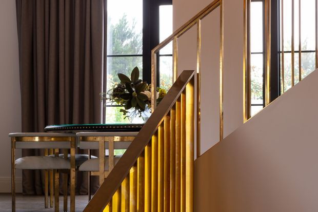 Interior view of a refined stairway with warm brass/gold vertical balusters and handrail, white walls, dark timber-toned stair elements, and tall black-framed windows. A small dining or console table with upholstered stools sits beside the staircase, with curtains and indoor planting adding softness.