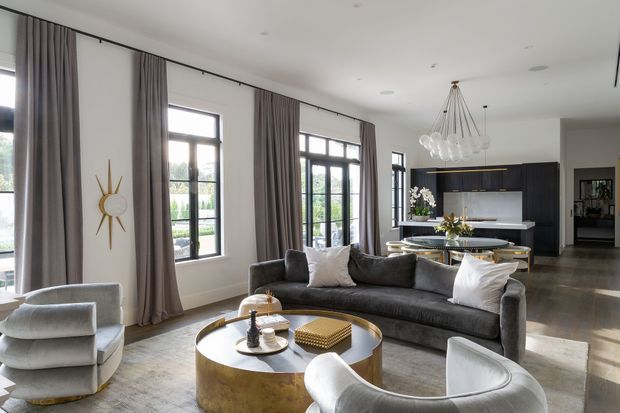 A refined open-plan living room with a large curved grey sectional sofa, sculptural light armchairs, a round metallic coffee table, tall black-framed windows dressed with full-height taupe curtains, and a contemporary black kitchen in the background. The space is bright, spacious, and elegantly styled, with a statement linear pendant chandelier and dark timber or stone flooring.