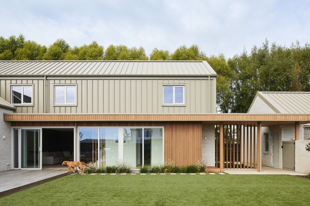 A contemporary two-storey residential extension with a long, low-pitched metal roof, pale vertical metal cladding on the upper level, and warm timber cladding along the ground-floor facade. Large glazed sliding doors open onto a timber deck and manicured lawn, while a timber slatted screen connects to the adjoining lower roof volume. Mature trees form the background.