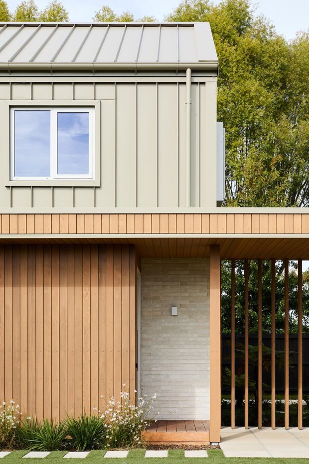 A close exterior view of a contemporary house featuring a pale grey-green standing-seam metal upper facade and roof, a projecting timber-clad lower level, and a sheltered entry porch. The entrance is framed by vertical timber posts and open timber screening, with a light brick feature wall and timber decking. Low planting and stepping stones border the paved approach.