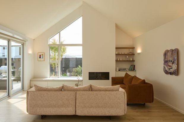 Warm, light-filled contemporary living room with a pitched ceiling, large floor-to-ceiling windows and glazed doors, pale timber flooring, and a built-in fireplace set into a central partition wall. A large neutral sectional sofa faces the fireplace, with a brown armchair, open timber shelving, wall lighting, and eclectic wall art completing the room.