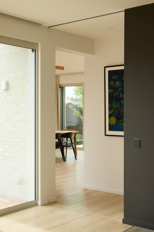 A warm, minimalist interior view looking through an open passage toward a dining area. The space features pale timber flooring, cream painted walls, a large glazed opening with a light brick exterior visible beyond, flush ceiling detailing, and a framed botanical artwork. A small timber dining table and dark chairs are visible in the adjoining room.