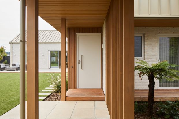 Covered residential entry porch with a modern white front door, timber-lined soffit, vertical timber cladding and substantial timber columns. Pale large-format tiled paving leads to the entrance, with a small lawn and stepping-stone path visible to the side. The adjacent facade combines light brickwork, dark-framed glazing and white upper cabinetry or screening.