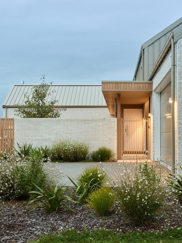 A contemporary residential entrance viewed from a landscaped front garden. The home features pale white brick walls, light grey standing-seam metal roofing and cladding, warm timber soffits and a timber slatted entry door beneath a projecting porch canopy. Native-style grasses and drought-tolerant planting surround a concrete path and paved entry court.