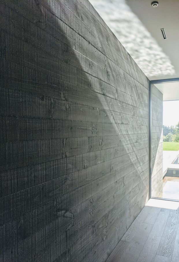 A minimalist interior corridor or passage with a dramatic dark textured concrete feature wall, wide grey timber-look flooring, and a large frameless glazed opening overlooking a bright outdoor area. The ceiling is white with recessed downlighting and a linear air vent, creating a strong contrast between the dark wall and daylight beyond.