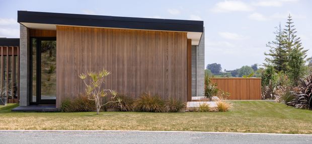 A single-storey contemporary building exterior with a strong horizontal flat roof, vertical timber wall cladding, dark metal fascia and window frames, and a contrasting pale masonry side wall. The front facade includes a recessed glazed entry and is set behind a neatly landscaped lawn with timber fencing and ornamental planting.