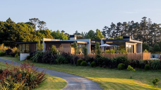 A single-storey contemporary residence set within a landscaped garden, viewed from a curving driveway. The house has a low horizontal composition with flat roof sections, dark cladding, large glazed openings, timber privacy screens and warm-toned timber fencing. Mature trees and dense ornamental planting surround the property.