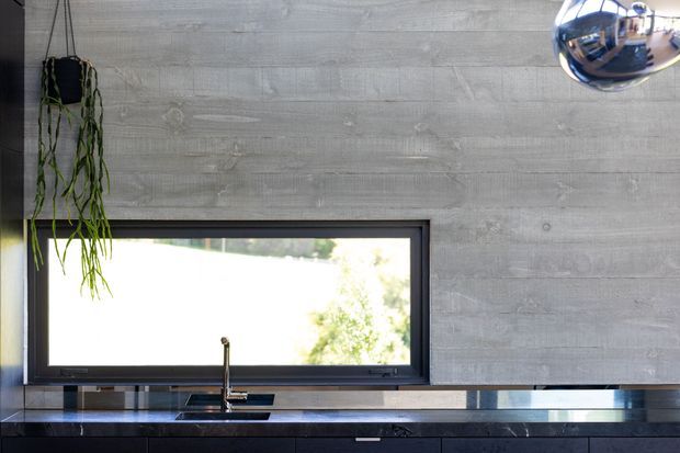Close-up view of a contemporary kitchen wall with a long horizontal black-framed window above a dark kitchen benchtop and undermount sink with a slim tap. The wall is finished in horizontally textured grey concrete-look panels, with dark lower cabinetry and a reflective chrome pendant light. A hanging plant is visible at the left edge, and greenery is seen through the window.