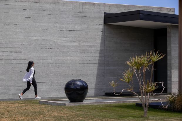 Exterior view of a minimalist contemporary residence featuring a broad textured concrete wall, a deep black projecting entrance canopy, dark recessed doorway, and a narrow paved threshold. A sculptural black planter and sparse architectural planting sit beside the entry, with a lawn in the foreground.
