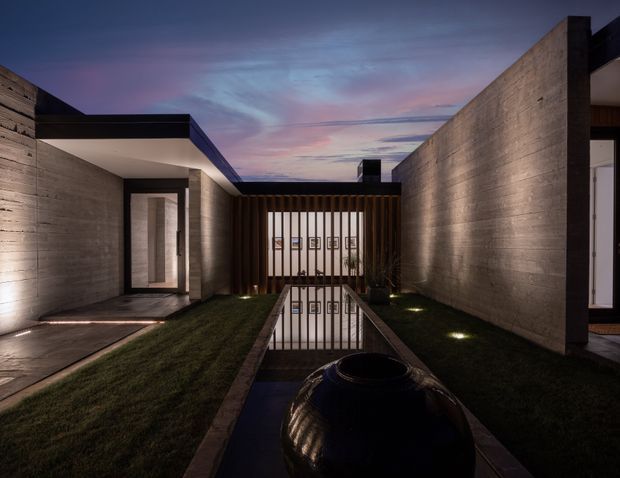 A dramatic dusk view of a modern architectural courtyard. Raw concrete walls form a narrow, symmetrical outdoor passage with strips of lawn, concealed ground lighting, and a long reflecting pool leading to a dark timber privacy screen. A large black urn or water feature sits in the foreground, while glazed openings connect to the building on either side. The flat-roofed composition and strong linear geometry create a calm, refined atmosphere.