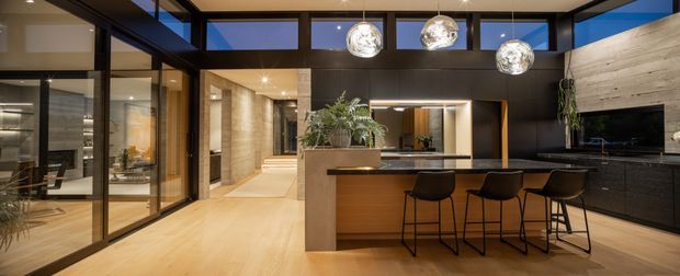 Open-plan contemporary kitchen and living area with a large island and three black bar stools. Floor-to-ceiling black-framed glazing and clerestory windows create a bright, connected space. The kitchen features dark built-in cabinetry, a warm timber island front, pale timber flooring, a textured light stone or concrete-look wall, and three spherical pendant lights. A living room with a wall-mounted television is visible to the right, while a dining or secondary seating area appears through the glazing on the left.