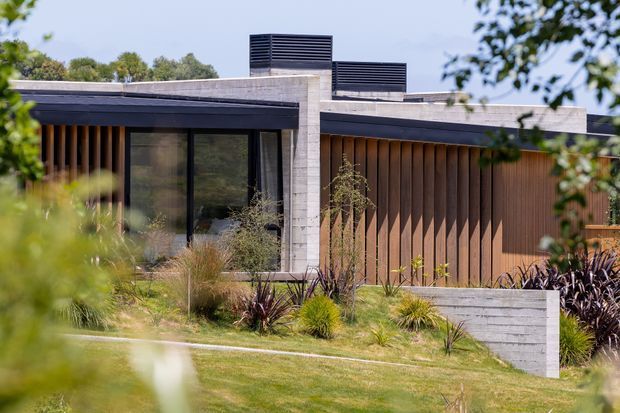 Exterior view of a low-profile contemporary single-storey building set within landscaped grounds. The facade combines horizontal concrete elements, dark metal-framed glazing, and vertical warm timber cladding, with flat roof planes and rooftop ventilation structures. Native-style ornamental planting and a gently sloping lawn frame the building.